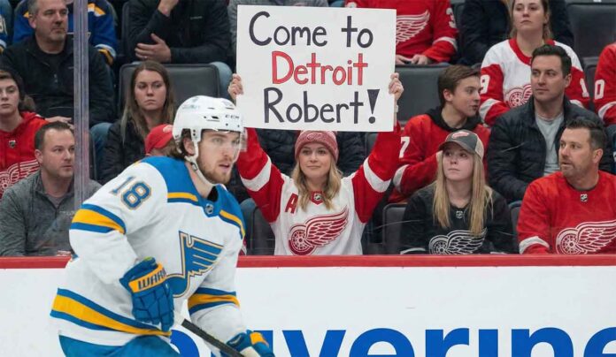 St. Louis Blues center Robert Thomas (18) skates on the ice during a game, while a female fan in a Detroit Red Wings jersey holds up a sign in the background that reads "Come to Detroit Robert!".
