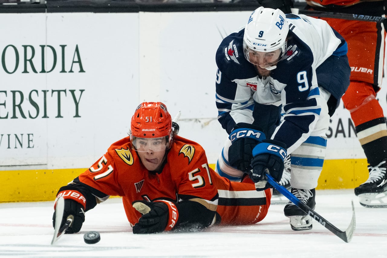 A Ducks player sticks his tongue out as he reaches for the puck while sliding on his stomach.