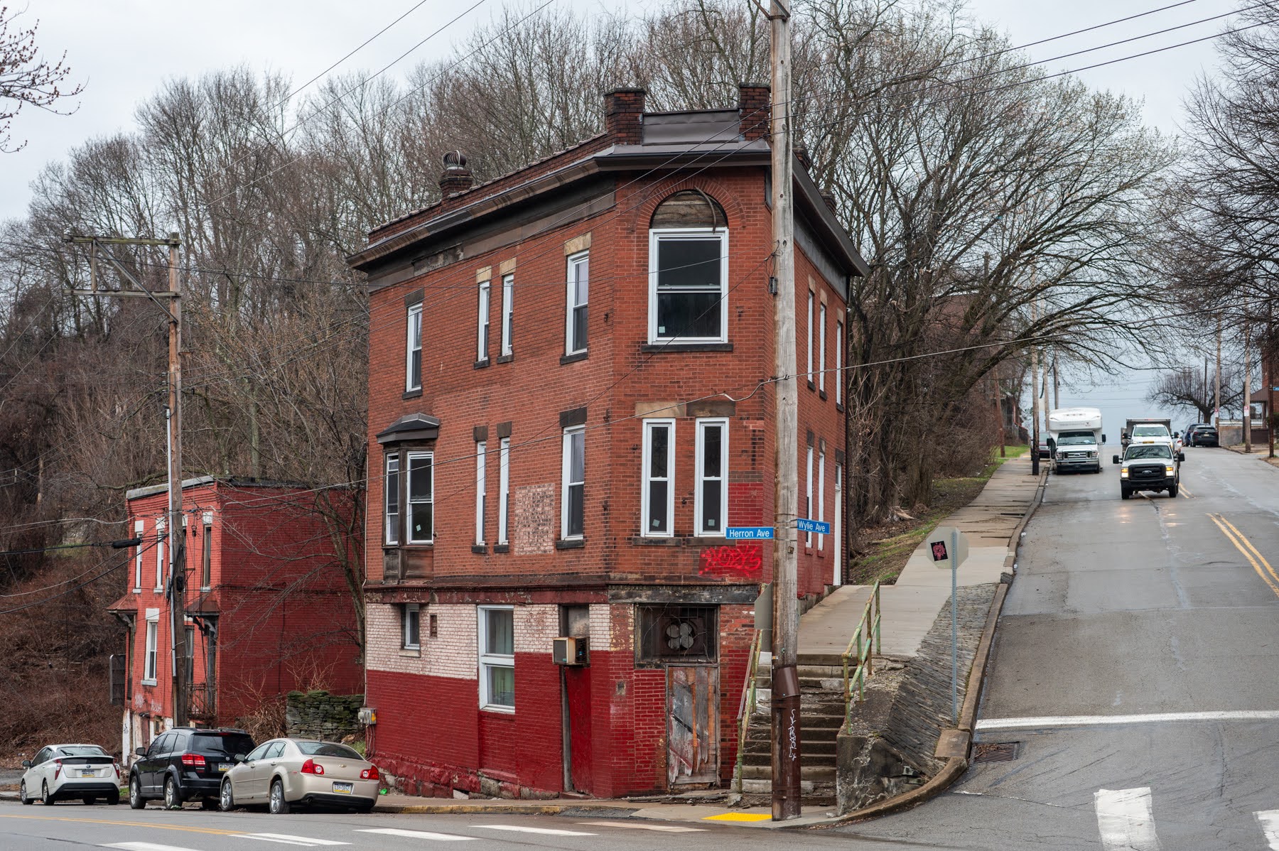 A worn, two-story brick building with boarded windows stands at a street corner on a cloudy day, with parked cars and trees in the background.