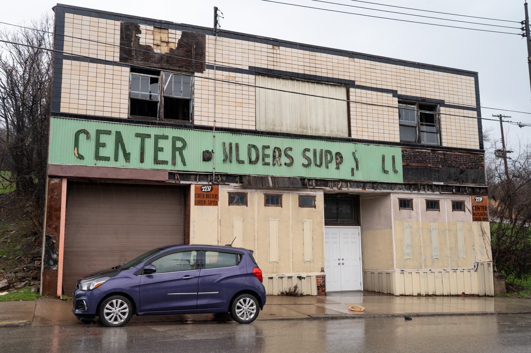 A damaged, abandoned two-story building with boarded windows and faded signage reading "CENTER BUILDERS SUPP LL." A purple car is parked in front on a wet street.