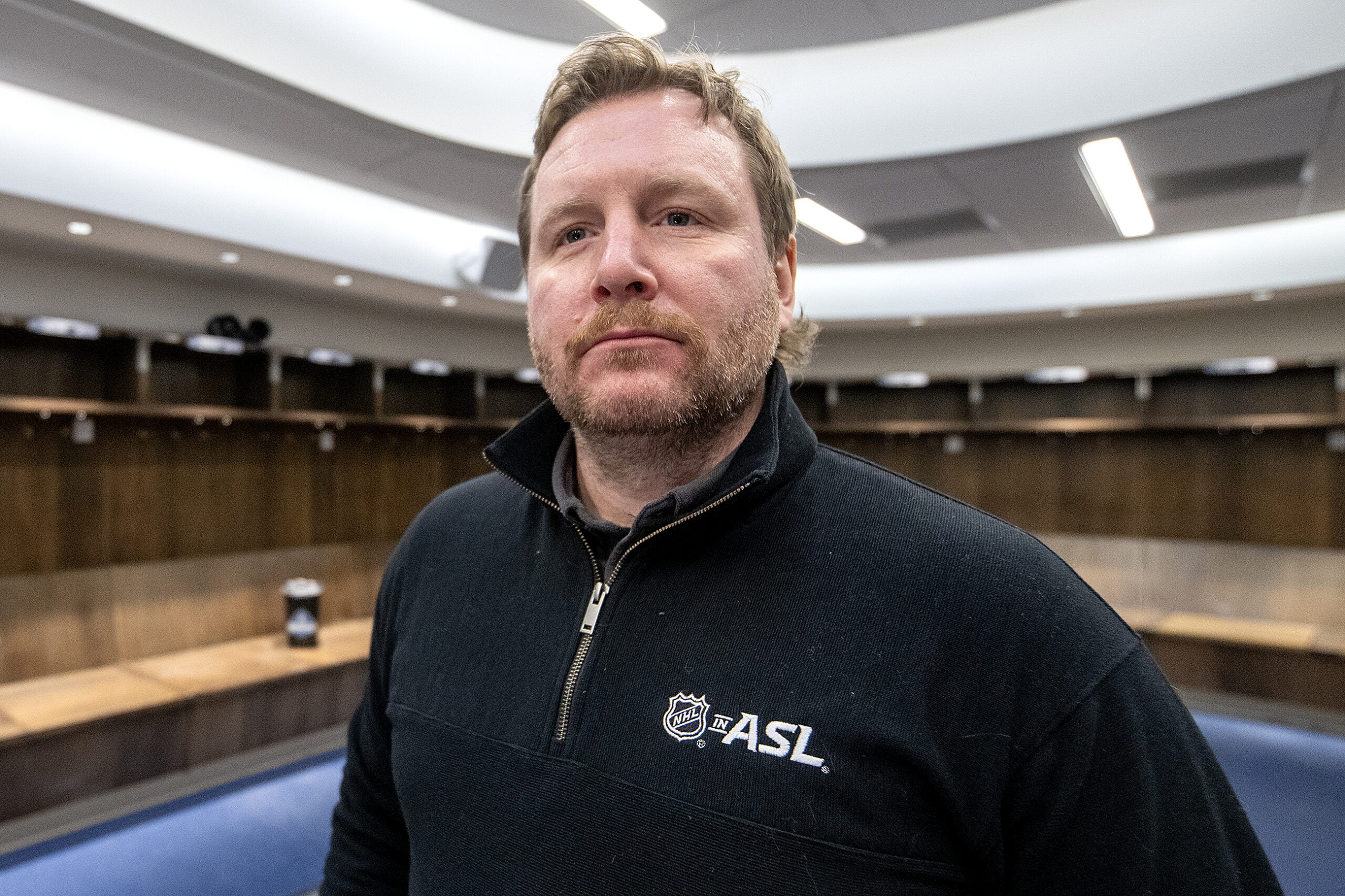 A man with light brown hair and a beard stands in an empty hockey locker room, wearing a black NHL ASL pullover.