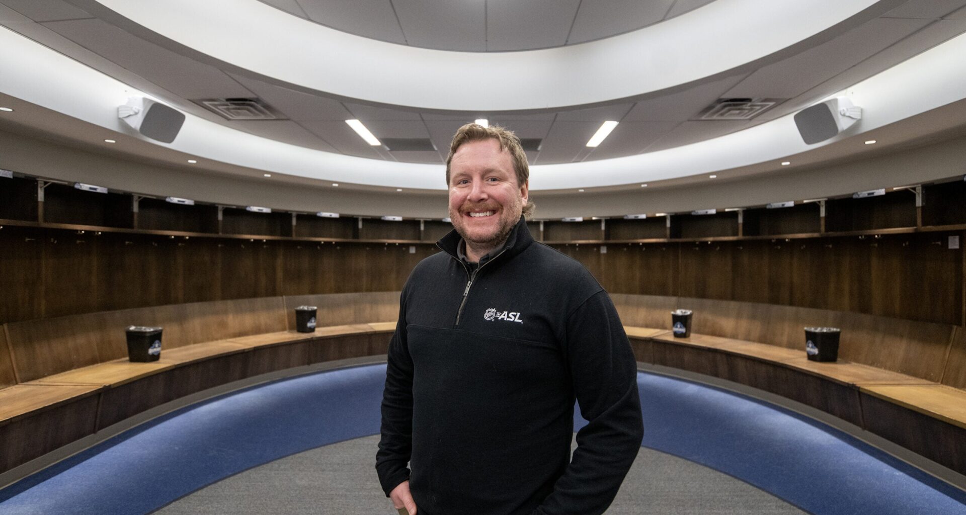 A man stands smiling in an empty, modern locker room with curved wooden benches and overhead lighting.