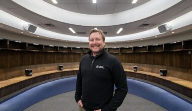 A man stands smiling in an empty, modern locker room with curved wooden benches and overhead lighting.