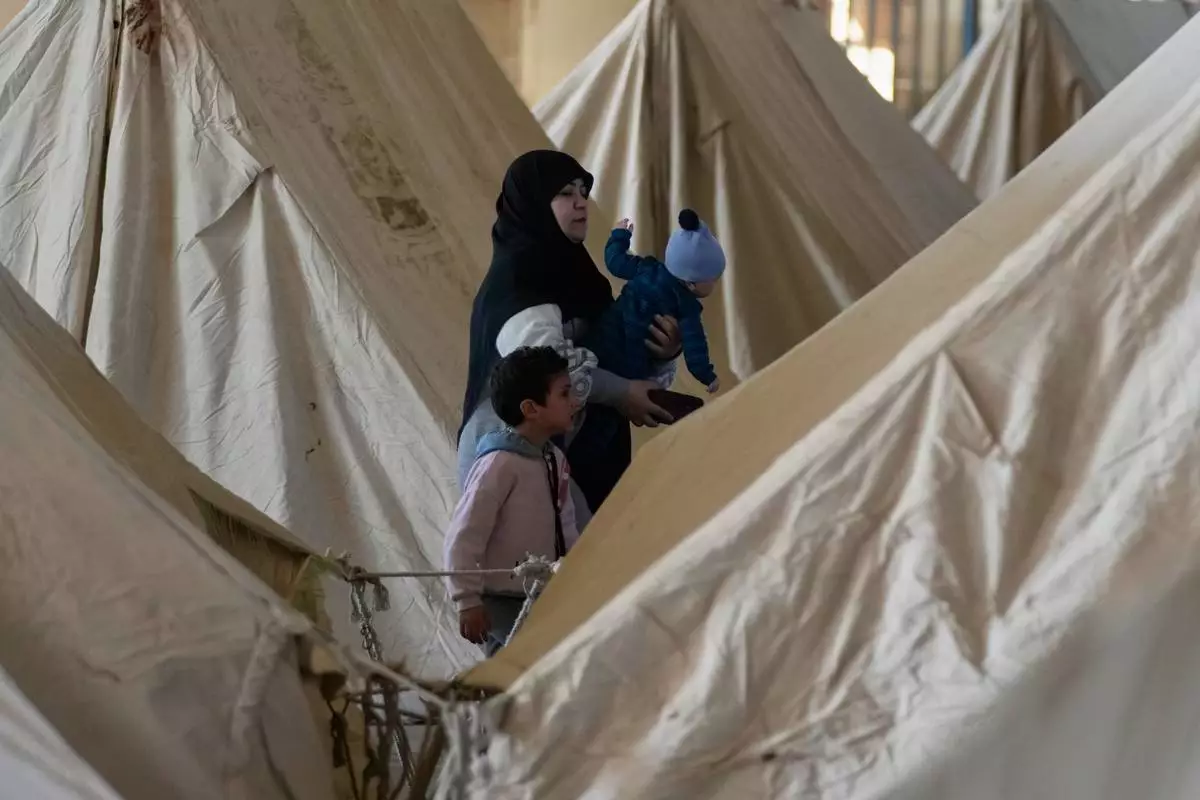 A displaced woman holds a child as another stands beside her between rows of tents at the Camille Chamoun Sports City Stadium, which has been turned into a shelter for people displaced by Israeli airstrikes in southern Lebanon and Dahiyeh, Beirut's southern suburbs, in Beirut, Lebanon, Tuesday, March 10, 2026. (AP Photo/Hassan Ammar)