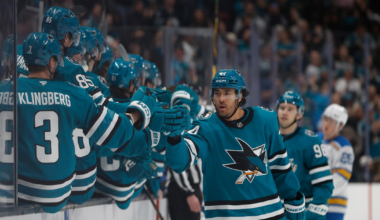 A hockey player in teal celebrates with teammates on the bench, giving fist bumps during a game with a crowd visible in the background.