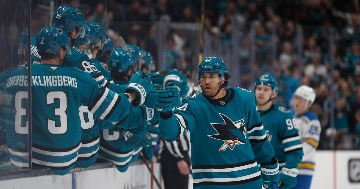 A hockey player in teal celebrates with teammates on the bench, giving fist bumps during a game with a crowd visible in the background.