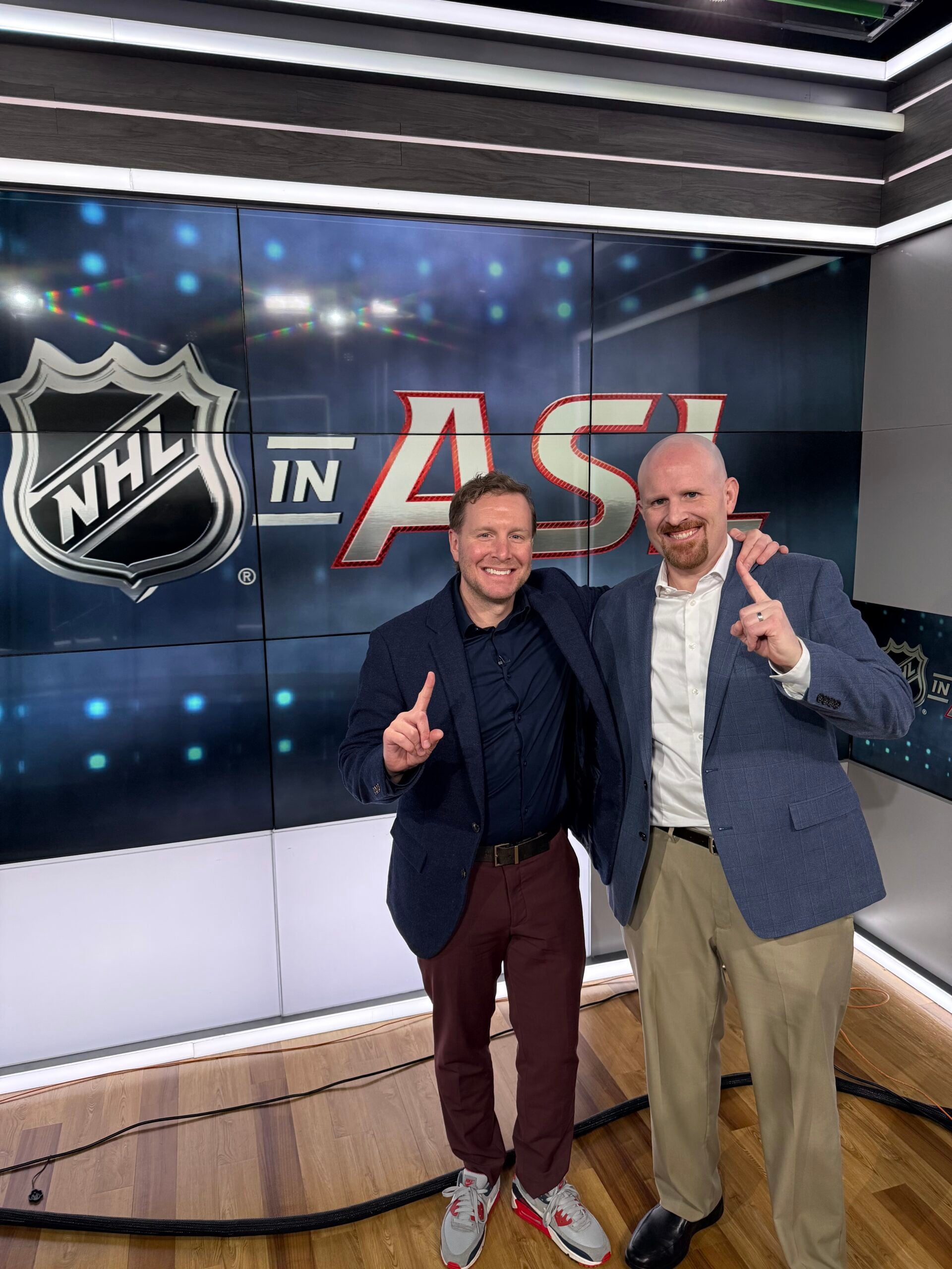 Two men in business attire stand smiling with raised index fingers in front of a display reading NHL in AS inside a studio.