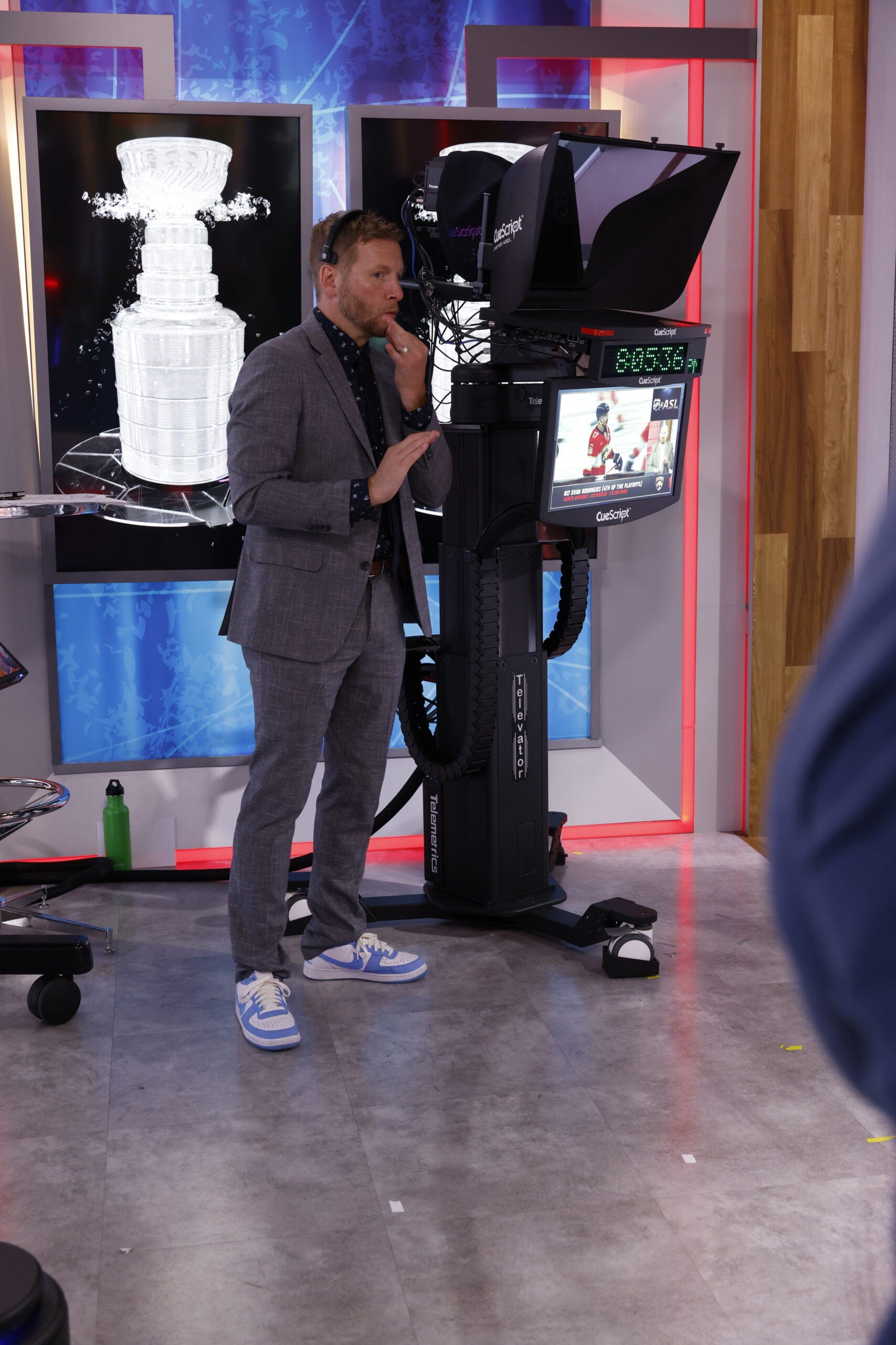 A man in a gray suit stands in a TV studio next to a camera and monitor, with an image of a hockey trophy displayed on a screen behind him.