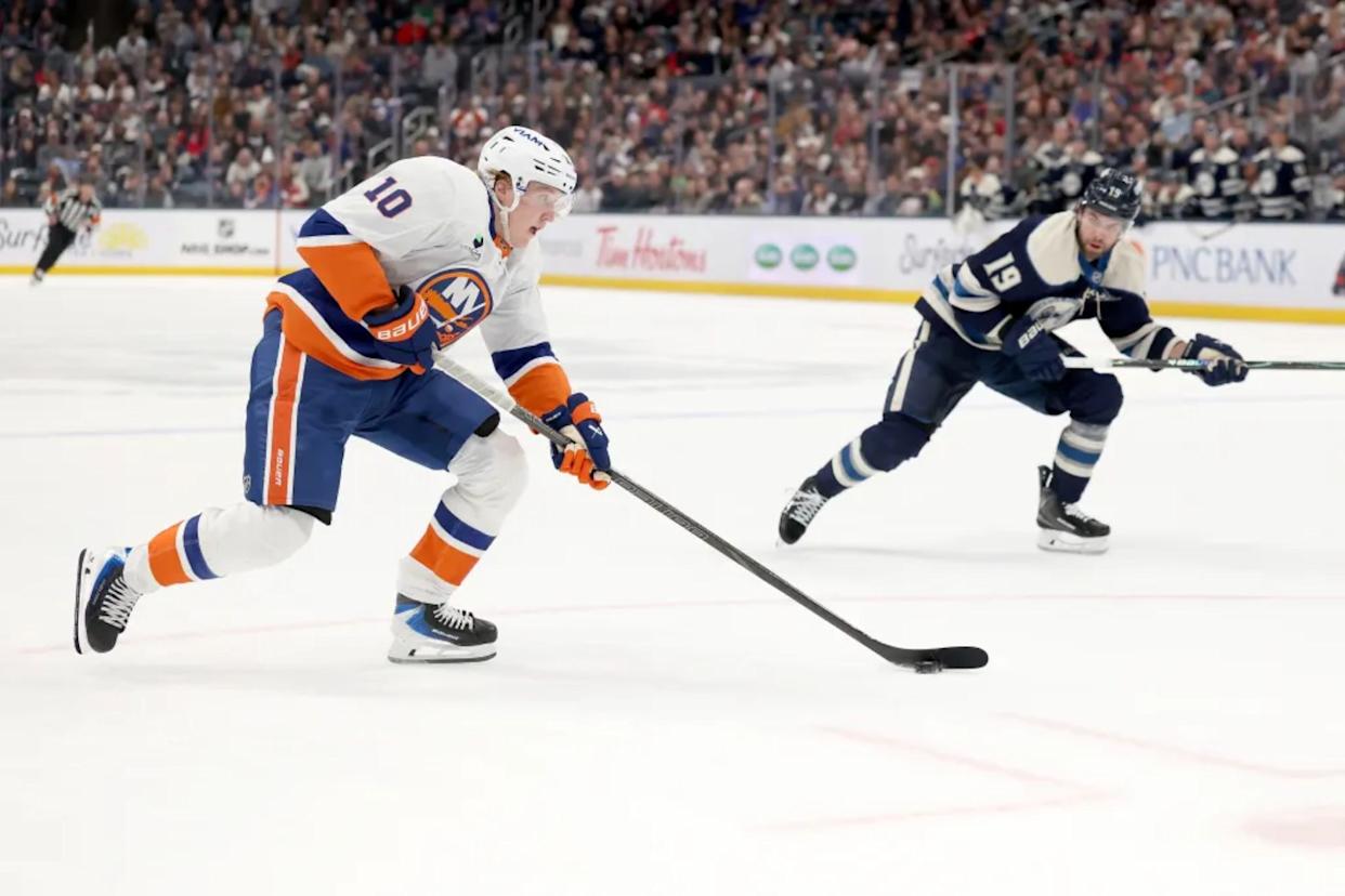 An image collage containing 1 images, Image 1 shows New York Islanders player Simon Holmstrom skating on ice with his hockey stick while Columbus Blue Jackets player Cole Sillinger skates behind him