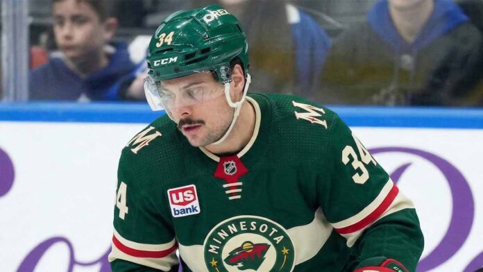 A close-up of Toronto Maple Leafs captain Auston Matthews (#34) in a Minnesota Wild jersey on the ice, appearing focused. Matthews is central to recent blockbuster NHL trade rumors linking him to the Wild.