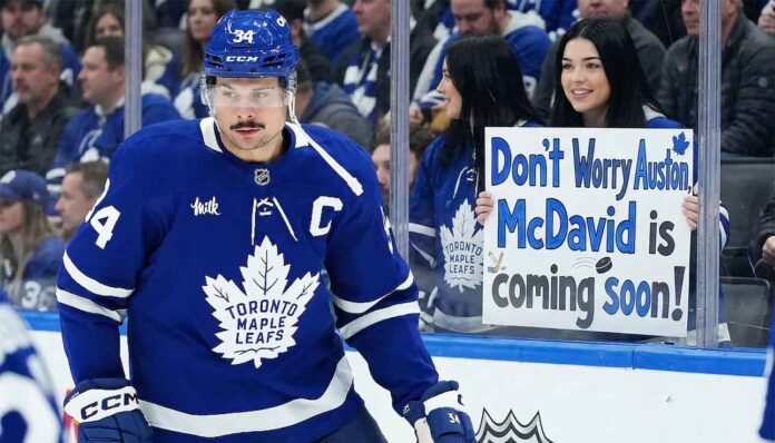 Toronto Maple Leafs captain Auston Matthews skates by the boards as a fan holds a controversial sign reading 'Don't Worry Auston, McDavid is coming soon!' highlighting fan frustration and future roster rumors.