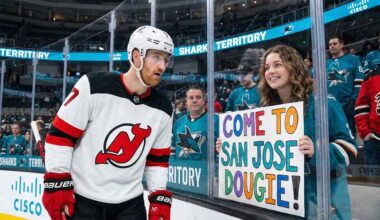 New Jersey Devils defenseman Dougie Hamilton looks at a hand-painted sign held by a smiling San Jose Sharks fan through the arena glass. The sign reads 'COME TO SAN JOSE DOUGIE!', referencing player movement and trade rumors between the two teams.