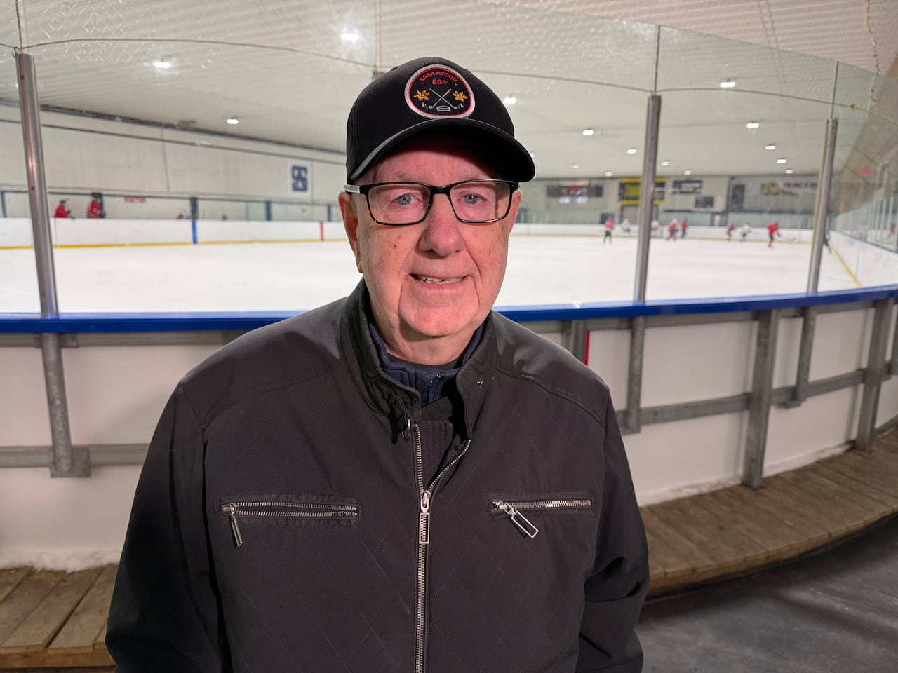A smiling, older man with glasses wearing a black jacket and a black ball camp stands in front of the ice surface in a hockey rink.