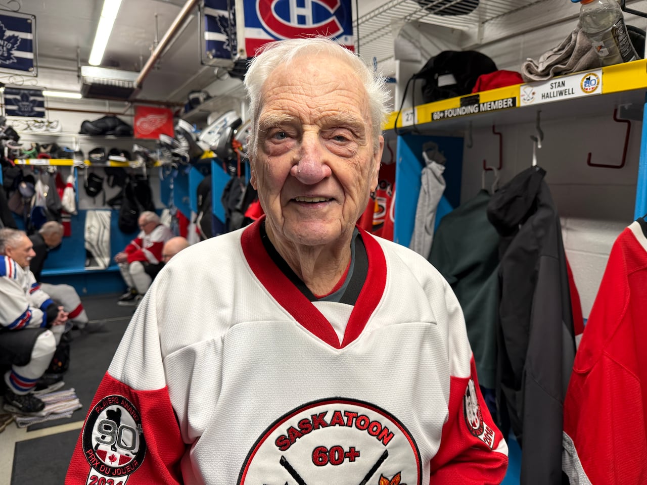 A smiling older man wearing a white and red hockey jersey stands in a hockey dressing room with other players, hockey equipment and NHL team banners in the background.