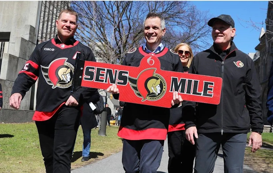  Accompanied by former Senators player Chris Neil, left, and team president Cyril Leeder, right, Mayor Mark Sutcliffe takes a Sens Mile street sign to a ceremony at City Hall on Friday. Sens Mile signs will be installed along Elgin Street for the length of the Senators’ run in the 2026 NHL playoffs.