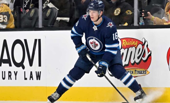 A close-up action shot on ice of hockey player Pavel Dorofeyev, featuring his likeness and low-crouched pose, now in a navy blue Winnipeg Jets home uniform and helmet with number 16, controlling a puck with his stick and creating ice spray.