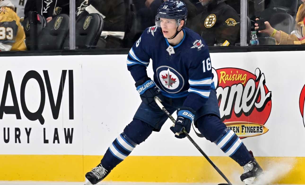 A close-up action shot on ice of hockey player Pavel Dorofeyev, featuring his likeness and low-crouched pose, now in a navy blue Winnipeg Jets home uniform and helmet with number 16, controlling a puck with his stick and creating ice spray.