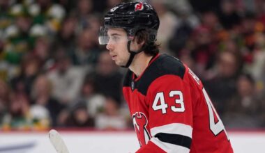 Close-up profile photograph of NHL star Quinn Hughes on the ice, wearing a New Jersey Devils home uniform and black helmet to visualize trade rumors that would reunite him with his brothers.