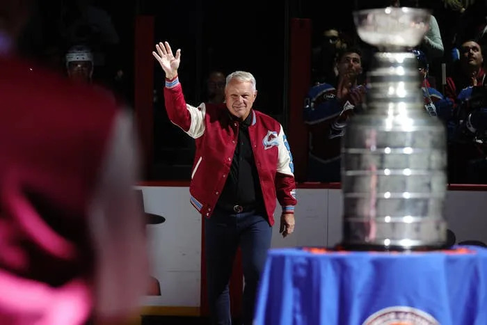 A person waves to the audience at a hockey event, wearing a varsity jacket, with a trophy on display in front