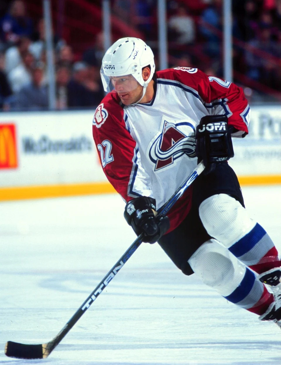 A hockey player in full gear, including a helmet and jersey, skates on the ice during a game, focused and determined. Arena background visible
