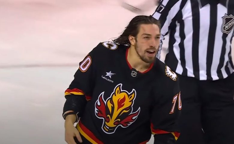Ryan Lomberg in a Calgary Flames alternate jersey (featuring a fiery horse logo) is shown on the ice during a game. His hair is wet and slicked back as he emerges from a heated altercation. An NHL referee is visible in the background.