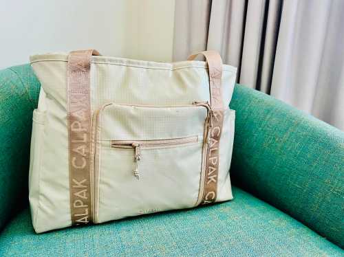 A light-colored tote bag with a front zipper pocket resting on a green couch.