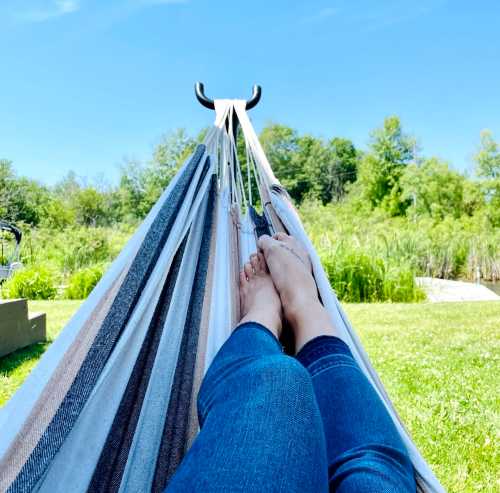 A person relaxing in a hammock with bare feet visible, surrounded by greenery and a clear blue sky.