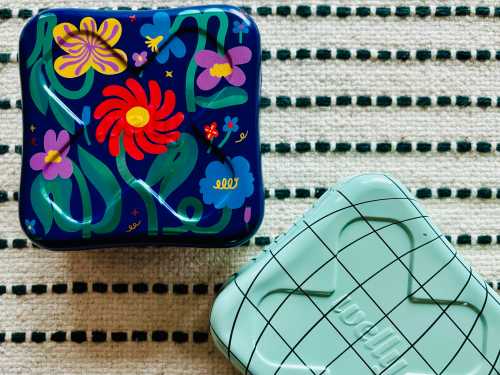 Two decorative metal tins with colorful floral designs, resting on a striped fabric surface.