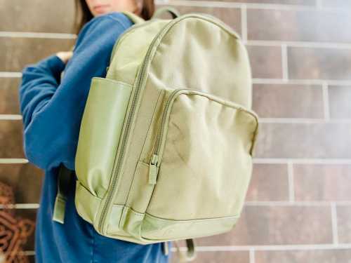 A person wearing a blue sweatshirt holds a green backpack against a textured wall.