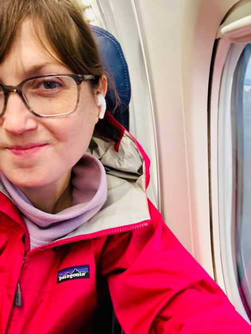 A person wearing glasses and a red jacket sits by an airplane window, smiling with earbuds in.