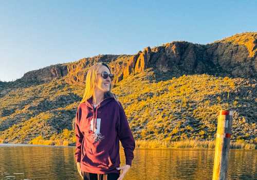 A person in a purple hoodie stands by a lake, with mountains and golden sunlight in the background.