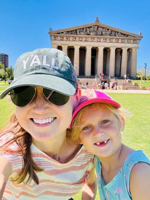 A woman and a young girl smile for a selfie in front of a large, classical-style building on a sunny day.