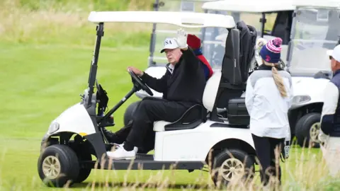 PA Media Donald Trump waves from a golf buggy while wearing dark-coloured waterproof golf clothes and a white cap. A woman wearing white waterproof jacket and blue wooly hat stands next to him