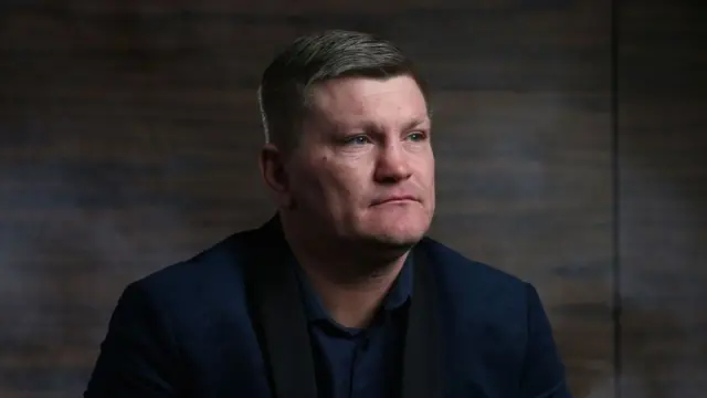 Close up of Ricky Hatton in a blue blazer and shirt sitting down in a dimly lit room with wooden-panelling walls