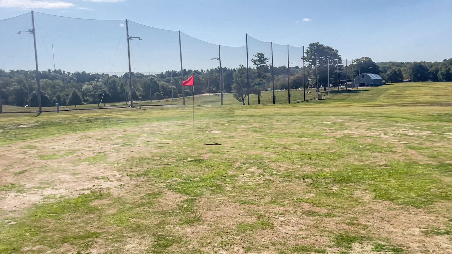 A wide area with patchy grass and dirt. There's a flag in a hole in the middle of the picture. In the background is a wall of netting bordering the driving range.