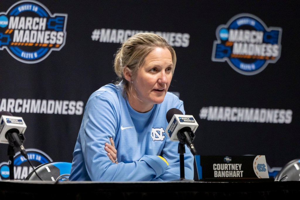 North Carolina head coach Courtney Banghart speaks with reporters ahead of an NCAA Tournament game. She is wearing a light blue pullover and leans forward in her chair with her arms across her chest.