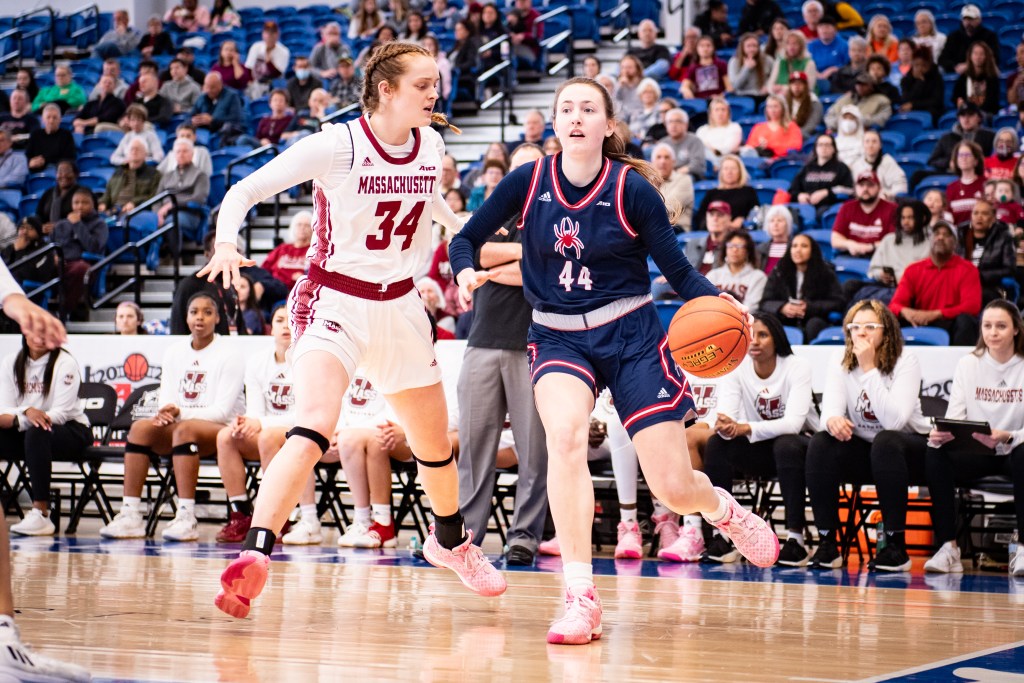 Richmond forward Maggie Doogan dribbles the ball with her left hand near the baseline. A UMass forward guards her tightly, cutting off her angle to get higher up the court.