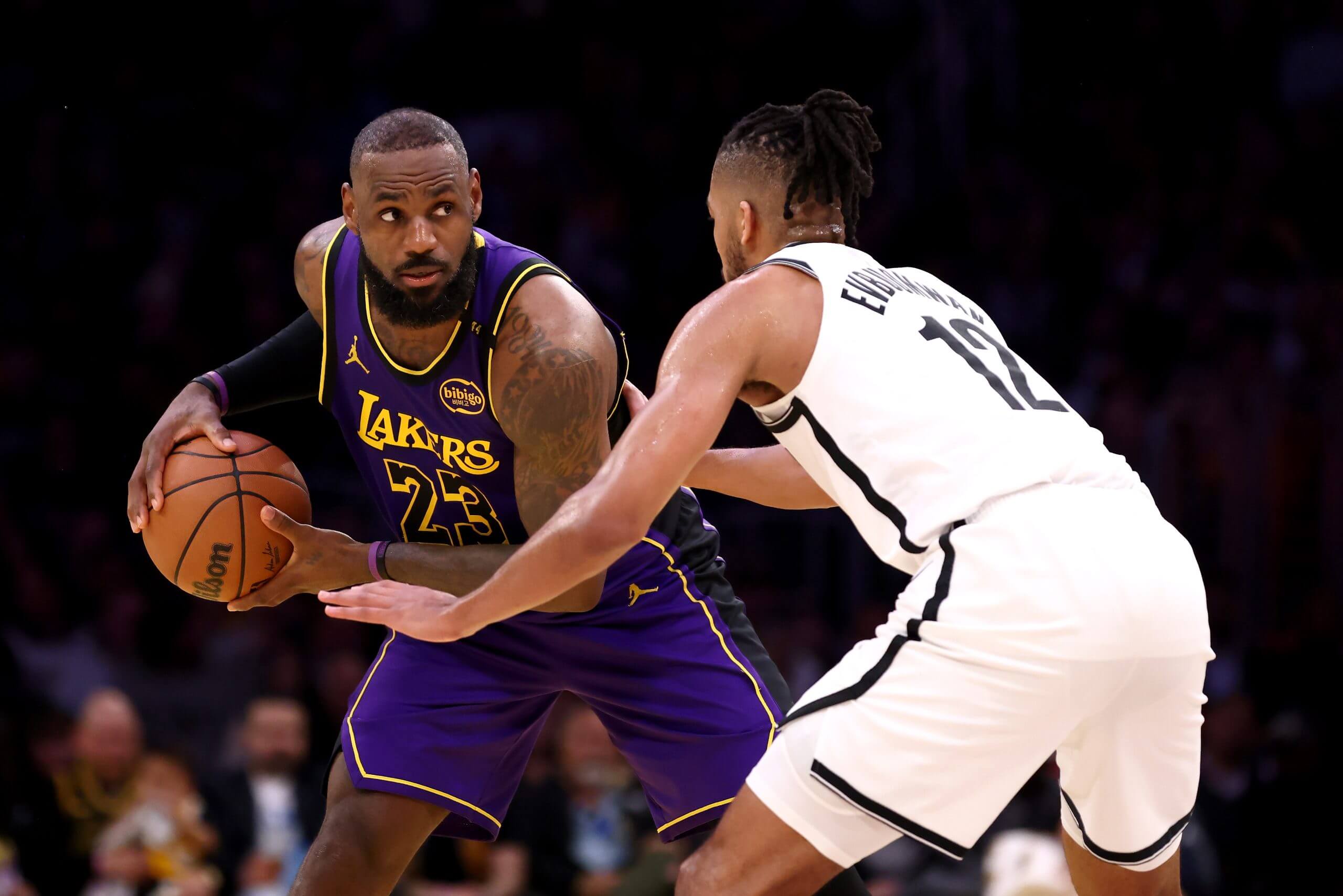 Tosan Evbuomwan, wearing a white Brooklyn Nets uniform, attempts to guard LA Lakers superstar LeBron James as she scans the court to make a pass
