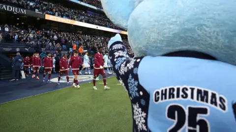 Getty Images City mascot Moonbeam in a blue Christmas jumper points to the teams as they walk out at the Etihad during the Premier League match between Manchester City and West Ham. 