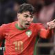 Brahim Diaz, visible from the waist up wearing a red Morocco shirt with green trim and the number 10 in gold on the front, screams in celebration and points his left hand in front of his body after scoring