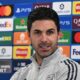 Mikel Arteta speaking at his news conference in a grey Arsenal tracksuit top, with a board of sponsor logos behind him