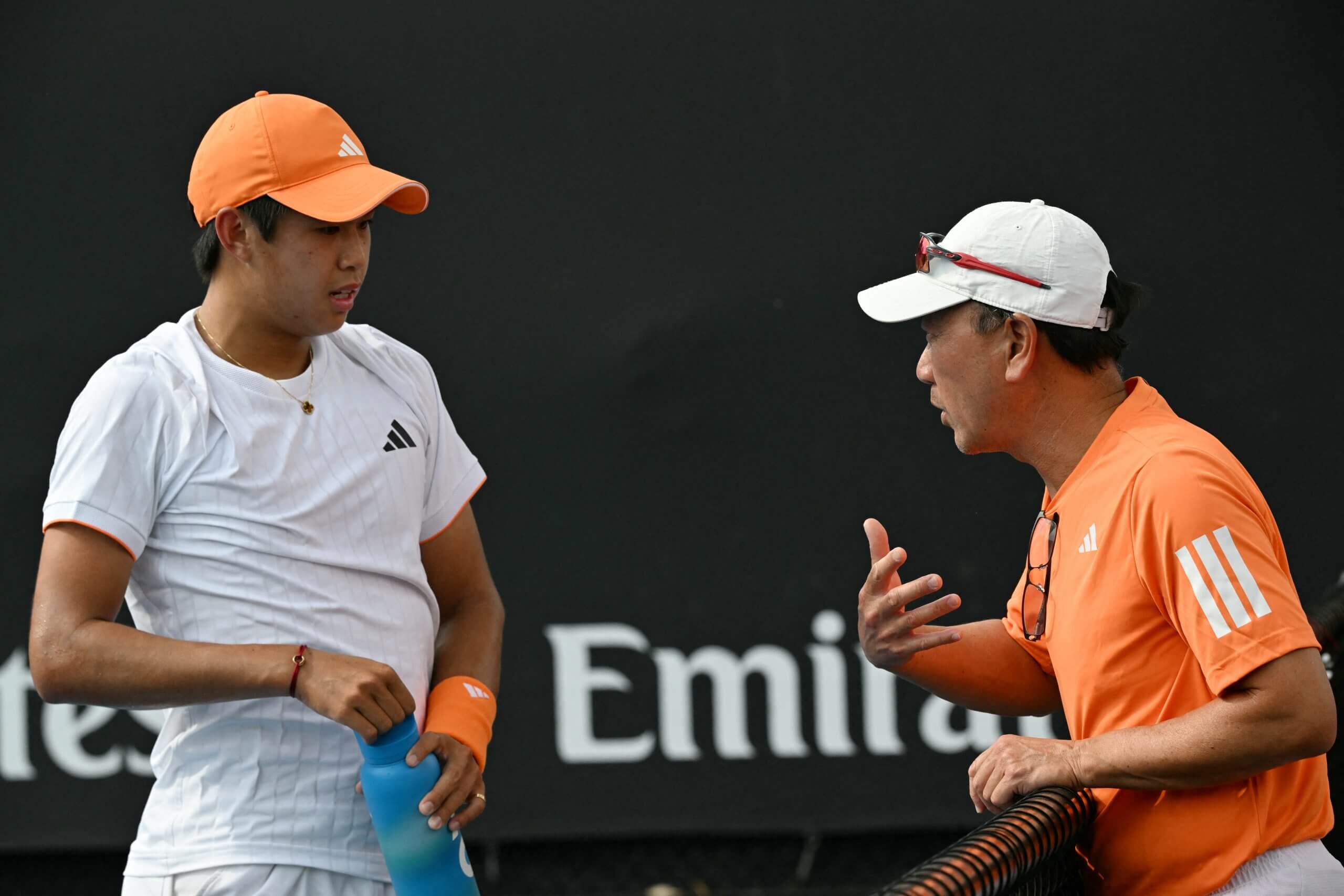 Learner Tien (left) wears a white shirt and orange cap and holds a water bottle as he talks to Michael Chang (right), who wears an orange shirt and gestures animatedly.