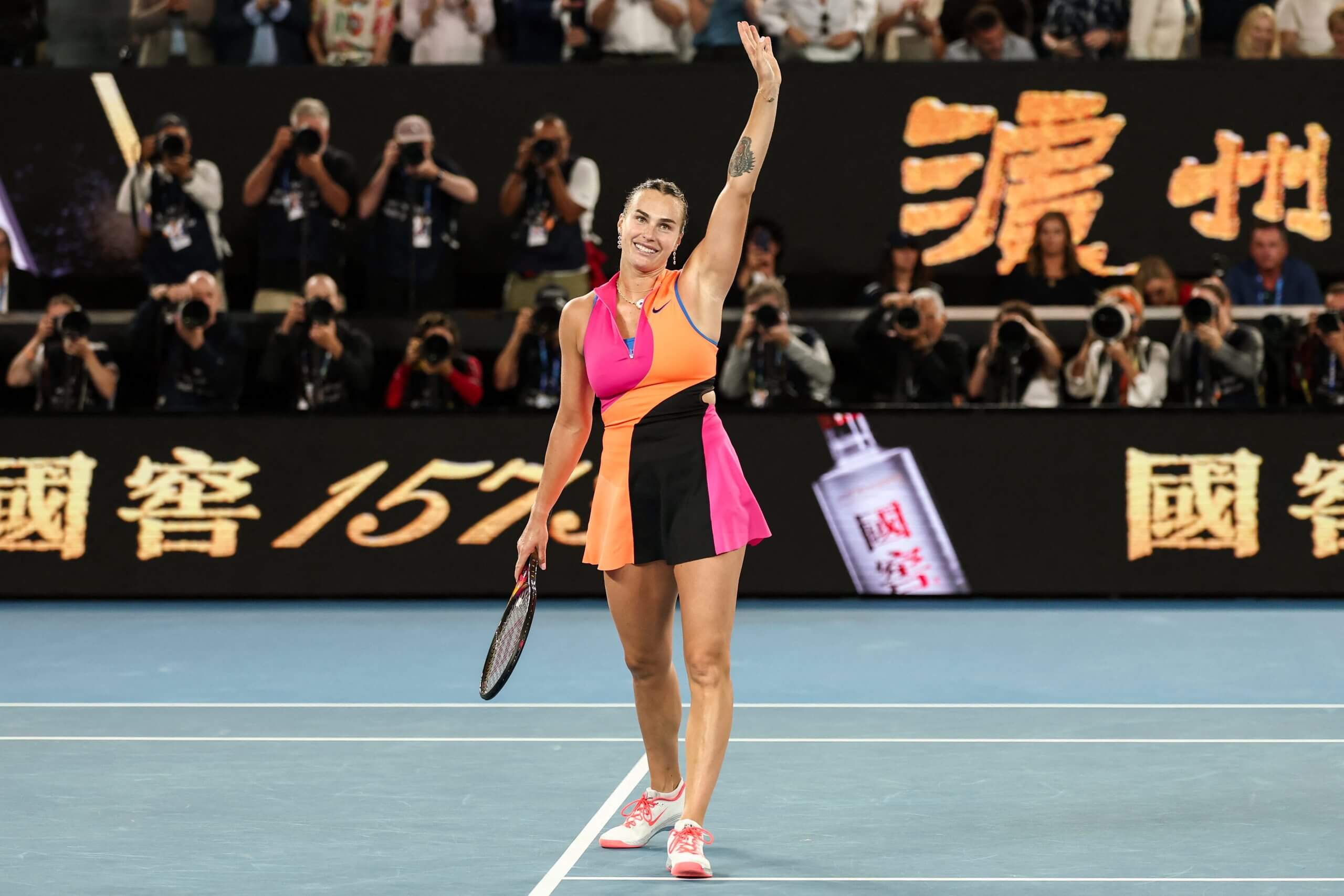 Aryna Sabalenka waves to the crowd wearing a pink, orange and black Nike kit.
