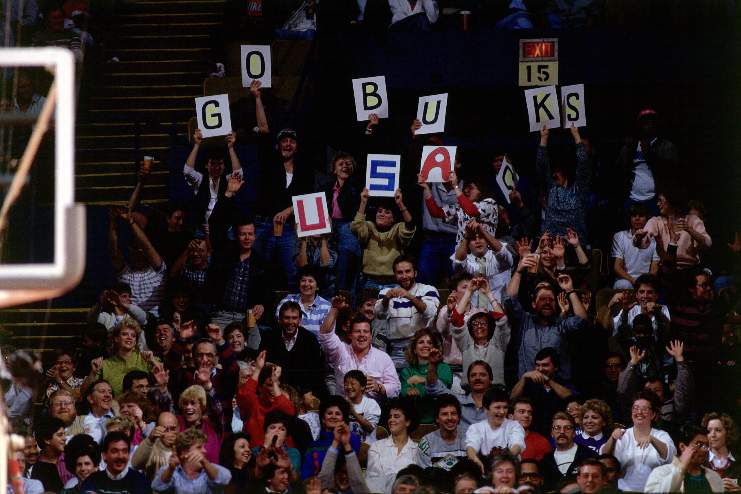 A crowd of sports fans in an arena hold up signs reading GO BUCKS and USA while cheering enthusiastically.