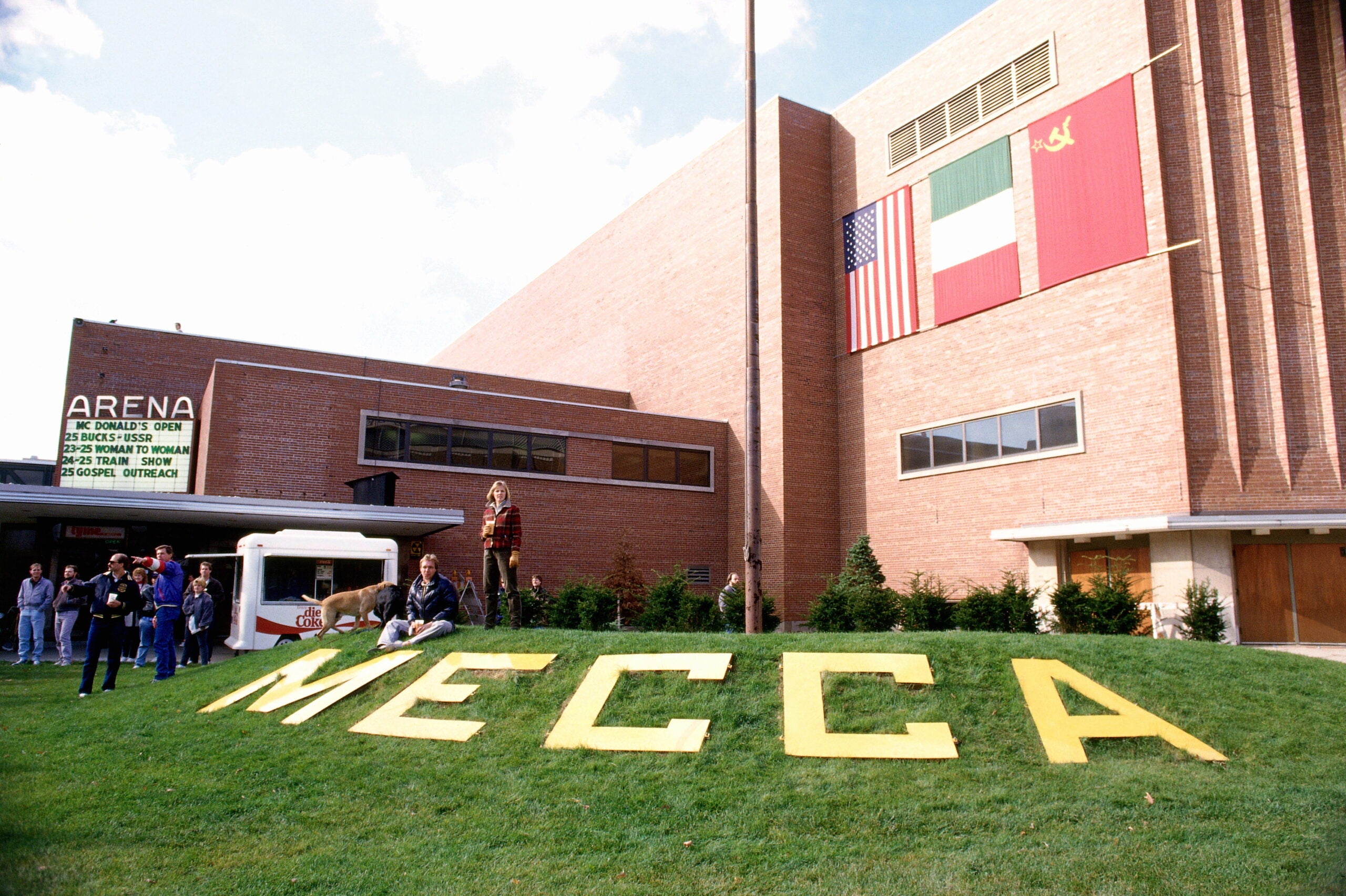 Group of people stand near a building with large yellow letters spelling MECCA on the lawn; international flags and a sign reading ARENA are visible in the background.