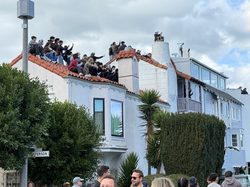 A large group of people sit closely together on a red-tiled rooftop, many holding up phones to capture photos or videos.