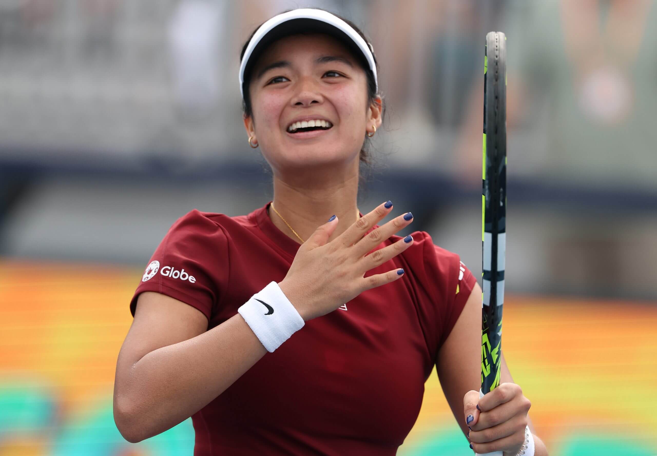 Alex Eala smiles widely as she holds her tennis racket in her left hand and places her right across her heart wearing a maroon tennis outfit.