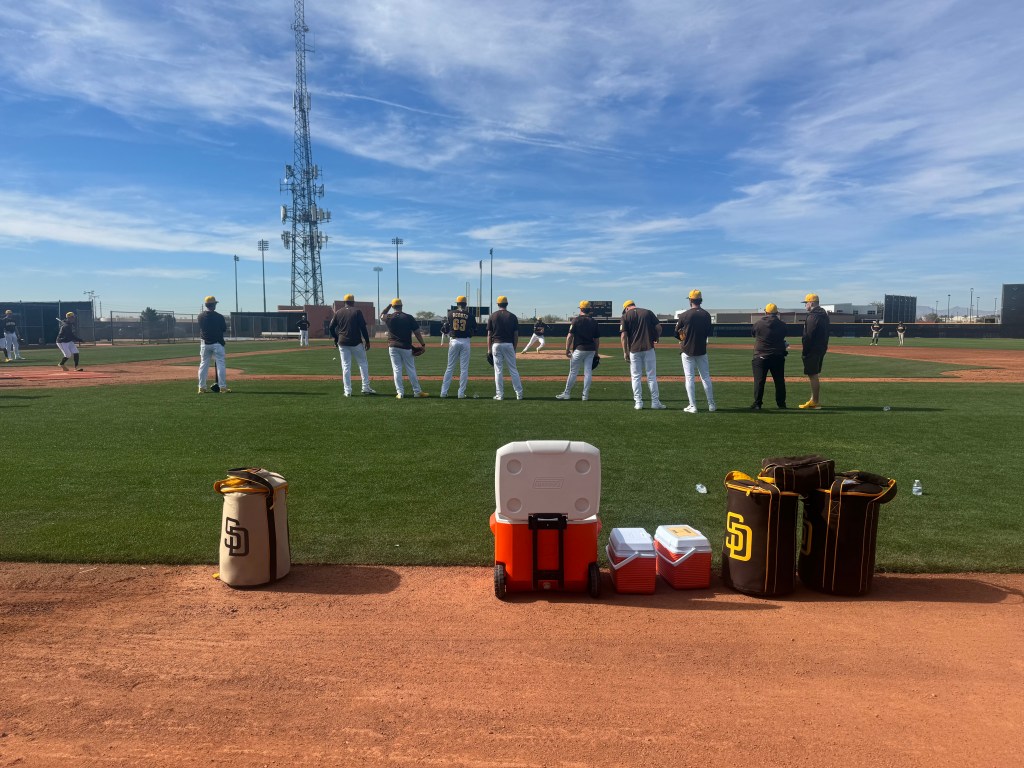 Coolers and other team-branded merchandise are placed on the dirt while coaches and players alike stand on the first base line.
