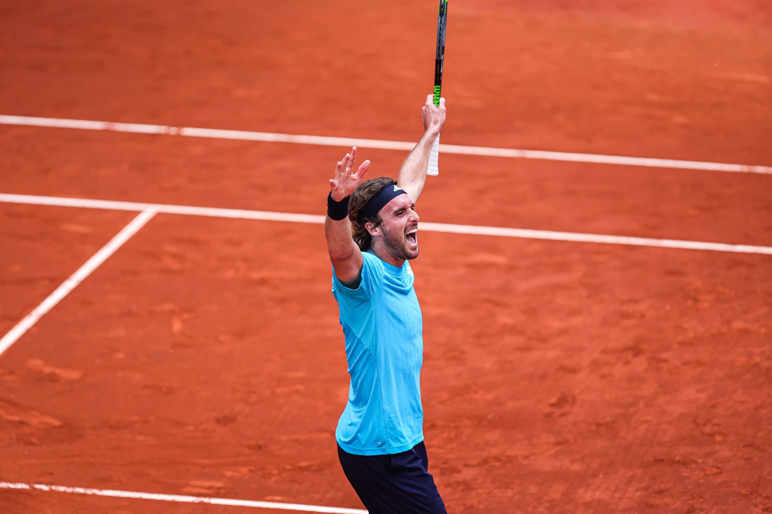 Stefanos Tsitsipas raises his arms aloft on a clay tennis court wearing a blue tennis shirt.