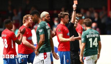 Paul Rutherford is shown a red card with his back to the camera showing his name and number in Wrexham's green away shirt, with Dagenham players in red shirts around the referee.